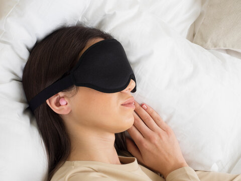 European Girl Getting Ready For Bed. A Pink Ear Plug And A Sleep Mask For Complete Darkness, Peace And Quiet All Night Long. Woman On A Pillow In The Bedroom. Healthy Lifestyle.