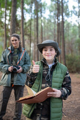 Fototapeta premium Joyful little boy with notebook in forest. Schoolboy in coat and panama smiling at camera, showing thumb up. Blurry mother in background. Childhood, nature, leisure concept