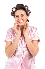 Happy young woman in pajamas and hair curlers on white background