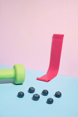 Trendy minimal sport concept with green dumbbell, booty band and blueberries on pink and blue...