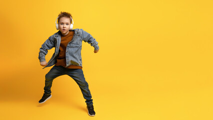 Fototapeta na wymiar full-length portrait of a 6-7-year-old kid in a denim shirt and a brown T-shirt with a hat on his head dancing with his arms outstretched, standing out against the pastel yellow background of child