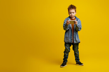 Cute kid schoolboy 6-7 years old playing on the phone, gadget, smartphone, isolated against a yellow wall, studio portrait. Concept of children of childhood, sale of gambling games for children