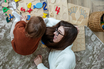 Mother and little daughter play educational games on rug in room. Toddler baby in a brown sweater.
