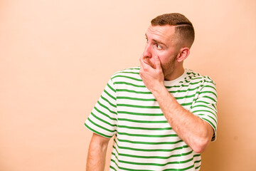 Young caucasian man isolated on beige background thoughtful looking to a copy space covering mouth with hand.