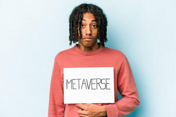 Young African American man holding metaverse placard isolated on blue background shrugs shoulders and open eyes confused.