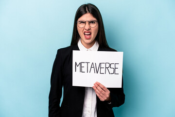 Young caucasian business woman holding a metaverse placard isolated on blue background screaming very angry and aggressive.