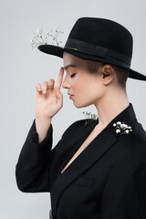 side view of woman with closed eyes pointing at black brim hat with gypsophila flowers isolated on grey.