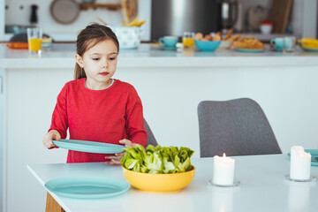 A young girl helps to set the family dining room table. Preschooler kid helping mother to set table in kitchen, carrying the plates, kitchen interior.