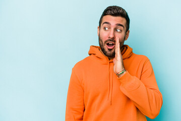 Young caucasian man isolated on blue background is saying a secret hot braking news and looking...