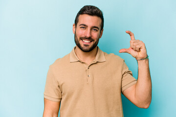 Young caucasian man isolated on blue background holding something little with forefingers, smiling...