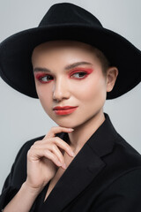 young woman with carmine red makeup, in black brim hat, touching chin isolated on grey.