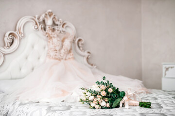 The bride's wedding bouquet of roses and green leaves lies on the bed, against the background of the bride's dress