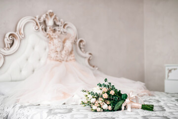The bride's wedding bouquet of roses and green leaves lies on the bed, against the background of the bride's dress