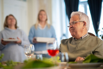 Smiling senior man sitting at dinner table with wife and adult daughter in background. Happy grandpa having dinner at home. Family party, event concept