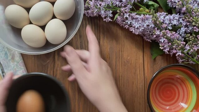 Women's hands take egg and break shell. Person separates egg white from yolk into different cups. Plate, flowers, decoupage tools on wooden table.