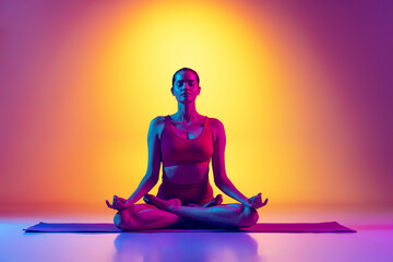 Young sportive woman sitting in lotus yoga pose on mat and doing meditation session isolated over gradient pink and yellow background in neon