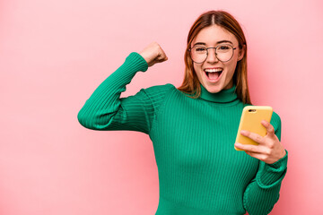 Young caucasian woman holding mobile phone isolated on pink background raising fist after a...