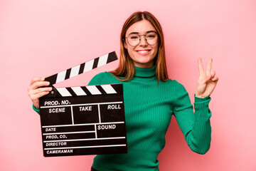 Young caucasian woman holding a clapperboard isolated on pink background showing number two with...