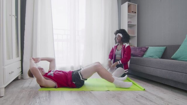 Funny retro style couple of Caucasian man and afro woman doing work up at home. Funny man doing abs exercises crunches at home with wife, fitness humor.