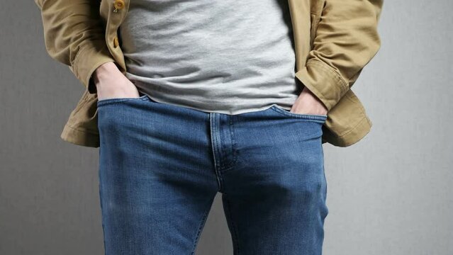 Poor man puts hands into jeans showing turned inside out empty pockets standing by grey wall. Male person looks for money in pants. Poverty concept closeup