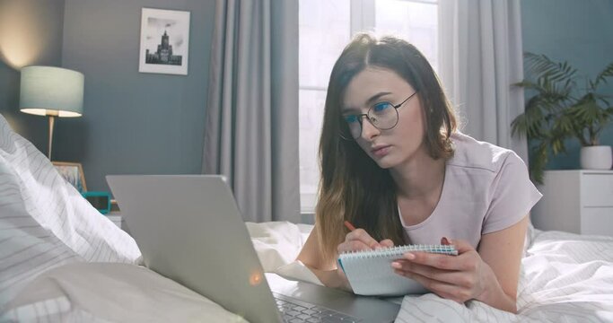 Young caucasian woman styding, making notes in paper notebook in bedroom. Pretty female wearing glasses, lying on bed and using laptop notebook to work.