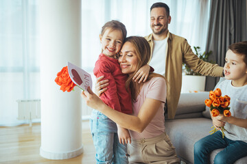 Smiling young woman reading greeting card while sitting on sofa with cheerful little siblings with bouquet of flowers during mother day. Happy mother getting gifts from kids
