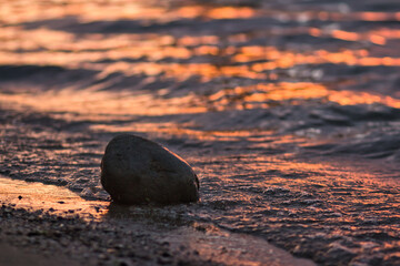 Close-up shot of a rock on the beach with reflecting sunset in the waves.
