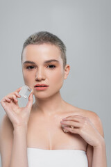 young woman with short hair holding ice cubes and looking at camera isolated on grey.