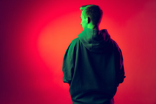 Back view portrait of young man in big stylish hoodie posing isolated over red studio background in green neon light - Powered by Adobe