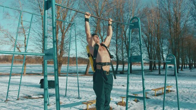 A muscular man with a naked torso with ballon drager on his back trains in a winter park and pulls himself up on a horizontal bar