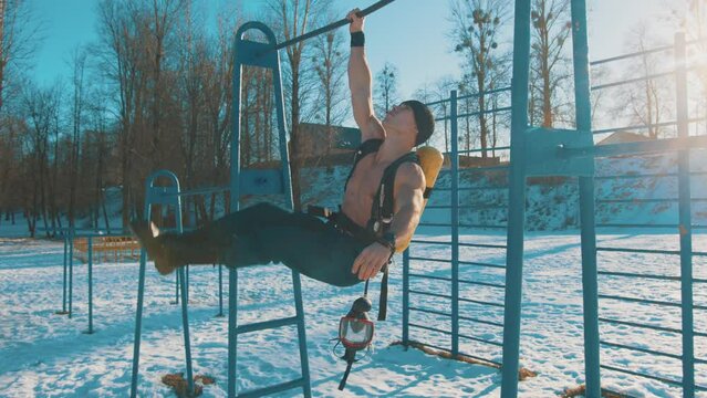 A bodybuilder without a T-shirt with a drager on his back hangs on the horizontal bar on one arm and raises his legs pumps up the press