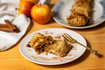 A roll of homemade baked fresh apple pie strudel with an appetizing slice of apple pie on the plate and fresh apples and cinnamon sticks on a wooden table. Rustic breakfast concept