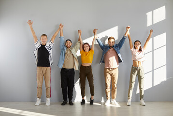 Obraz premium Happy people in casual clothes. Indoor studio group shot of young models in comfortable T shirts, denim jackets, pants, trousers, sneakers and trainers standing in row and holding and raising hands up