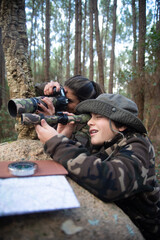 Pleased family taking pictures in forest. Mother and son with modern cameras lying on ground, peeking from stone. Parenting, family, leisure concept