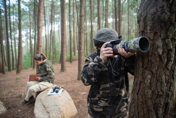 Fototapeta premium Absorbed boy with camera in forest. Schoolboy in coat and panama taking pictures. Blurry mother in background. Childhood, nature, leisure concept