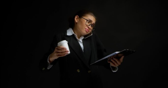 A busy Asian business woman in office clothes is holding coffee in white paper cup and documents in hands, talking discontentedly on the phone holding it with her shoulder.Isolated on black background