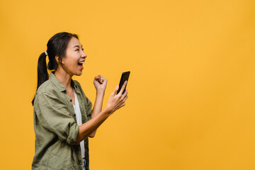 Surprised young Asia lady using mobile phone with positive expression, smiles broadly, dressed in casual clothing and stand isolated on yellow background. Happy adorable glad woman rejoices success.