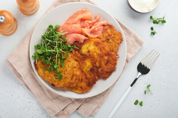 Potato pancakes. Fried homemade potato pancakes or latkes with cream, green onions, microgreens, red salmon and sauce in rustic plate on white table background. Rustic style. Healthy food. Top view.