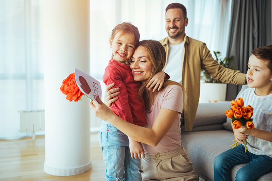 Smiling young woman reading greeting card while sitting on sofa with cheerful little siblings with bouquet of flowers during mother day. Happy mother getting gifts from kids