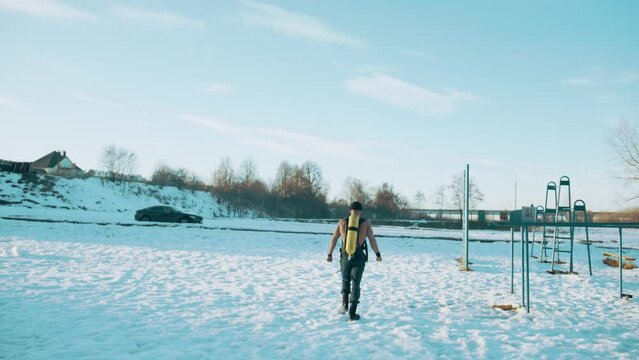 A muscular man without a T-shirt with a drager on his back goes through the snow to the horizontal bars. Back view