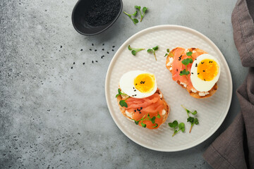 Sandwich with delicious toppings smoked salmon, eggs, herbs and microgreens radish, black sesame seeds over white plate on gray concrete table background. Healthy open sandwich superfood. Top view.