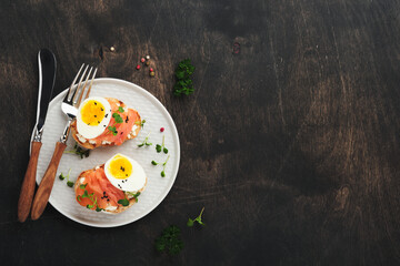 Sandwich with delicious toppings smoked salmon, eggs, herbs and microgreens radish, black sesame seeds over white plate on old wooden table background. Healthy open sandwich superfood. Top view.