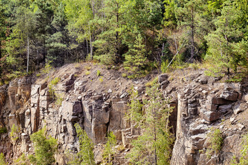 Rocky slope of quarry with sharp stones overgrown with trees. Abandoned granite quarry. Summer landscape with cliffs and cloudy sky.