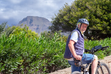 Happy active senior man with electro bikes standing outdoors in the park. Smiling elderly...