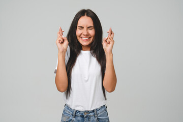 Hoping caucasian young woman in white T-shirt crossing fingers for good luck, success, winning money on bet lottery online casino isolated in grey background