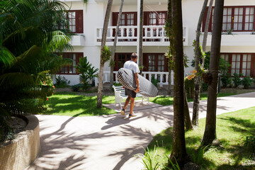 Naklejka na ściany i meble Man goes with surfboard in hotel. Back view. Active lifestyle. Tropical environment.