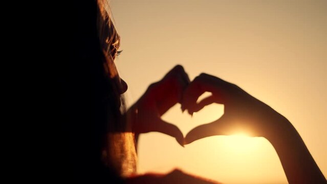 Happy girl in park at sunset. Finger-shaped heart shape. Hands of girl shape of heart. Summer dream. Happiness of freedom in a field at sunset.Sunlight between fingers.Silhouette of happy girl in park