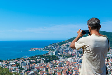 Fototapeta na wymiar Man tourist takes a photo of the architecture Budva city from a hilltop. Panoramic landscape. Top view. Adriatic sea. Montenegro