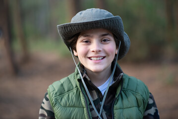 Portrait of cute boy in panama in forest. Dark-haired schoolboy in coat smiling at camera. Childhood, nature, leisure concept