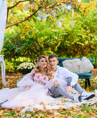 Front view of beautiful wife and husband, wearing in fashionable clothes, sitting together on carpet on grass against of trees and plants background, smiling and looking at camera in autumn day